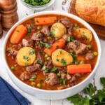 A bowl of lamb stew with carrots, potatoes, and peas, next to bread.