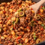 A wooden spoon scooping picadillo from a skillet, featuring meat, olives, and veggies.