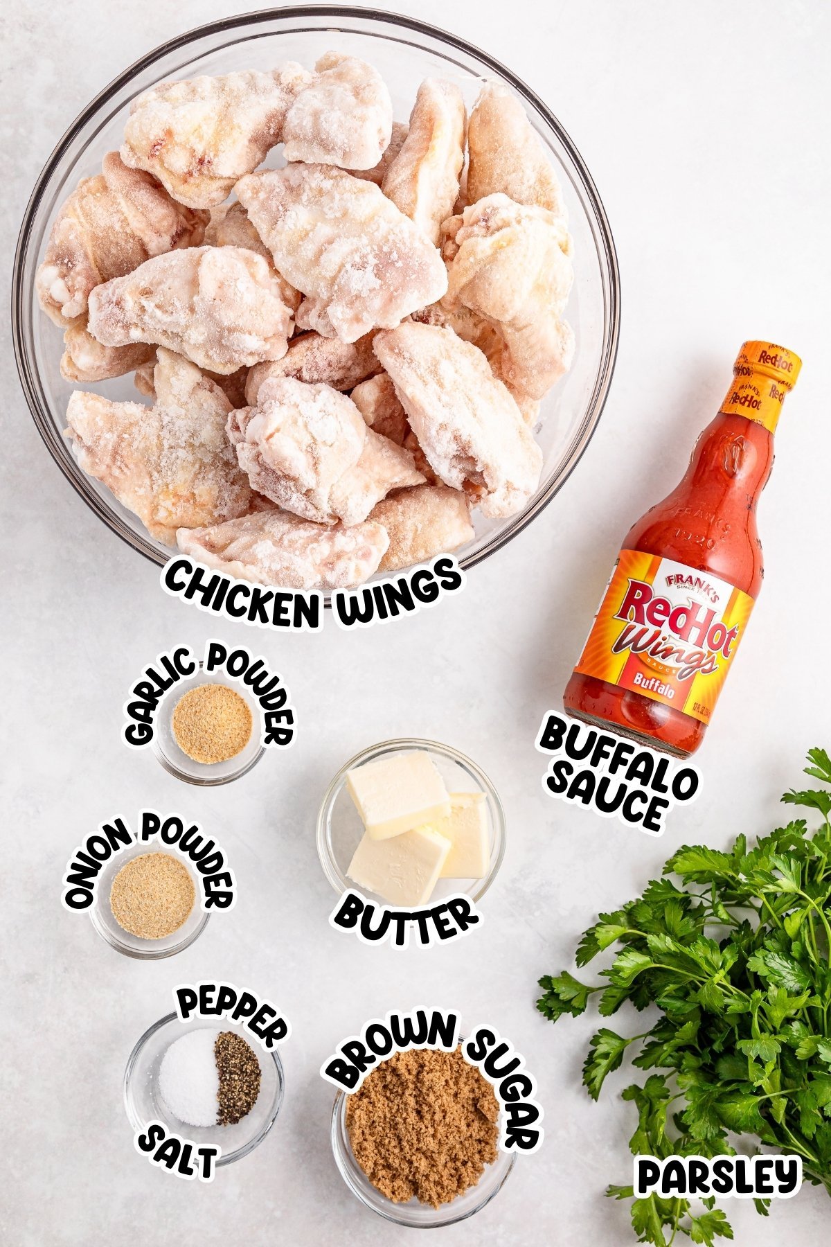 Frozen chicken wings, buffalo sauce, spices, butter, and parsley on a white surface.