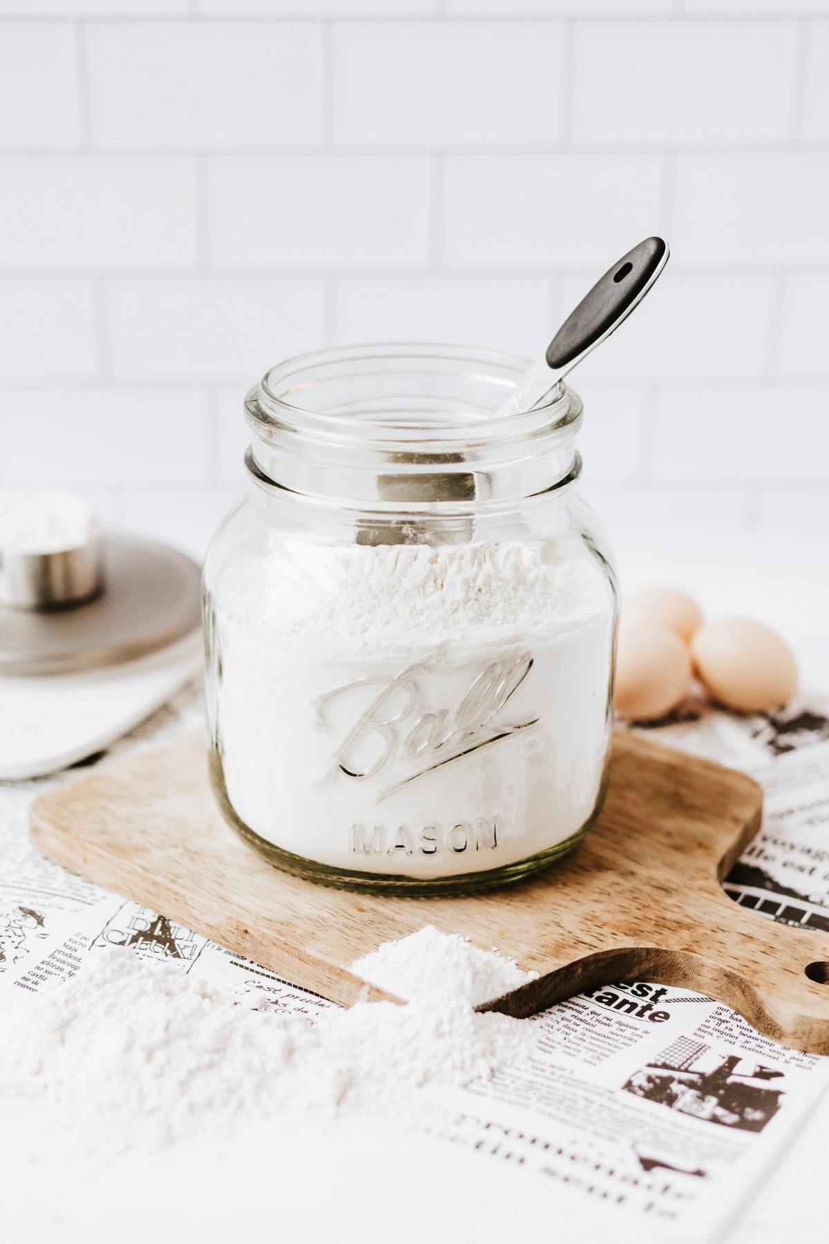Jar of flour with a measuring spoon on a wooden board, eggs nearby.