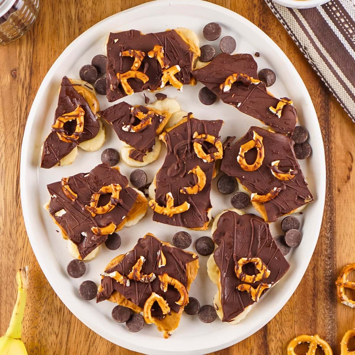 Chocolate-covered banana slices with pretzels on a white plate.