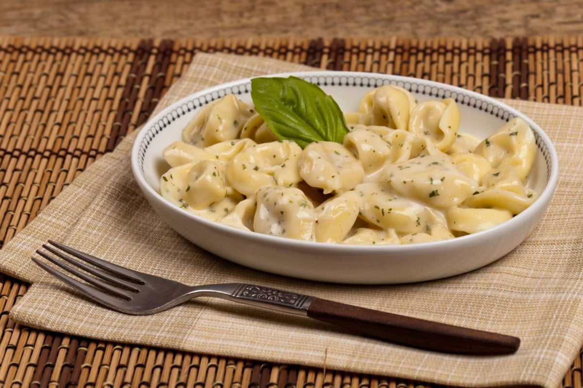 A bowl of creamy tortellini pasta garnished with a basil leaf, beside a fork.