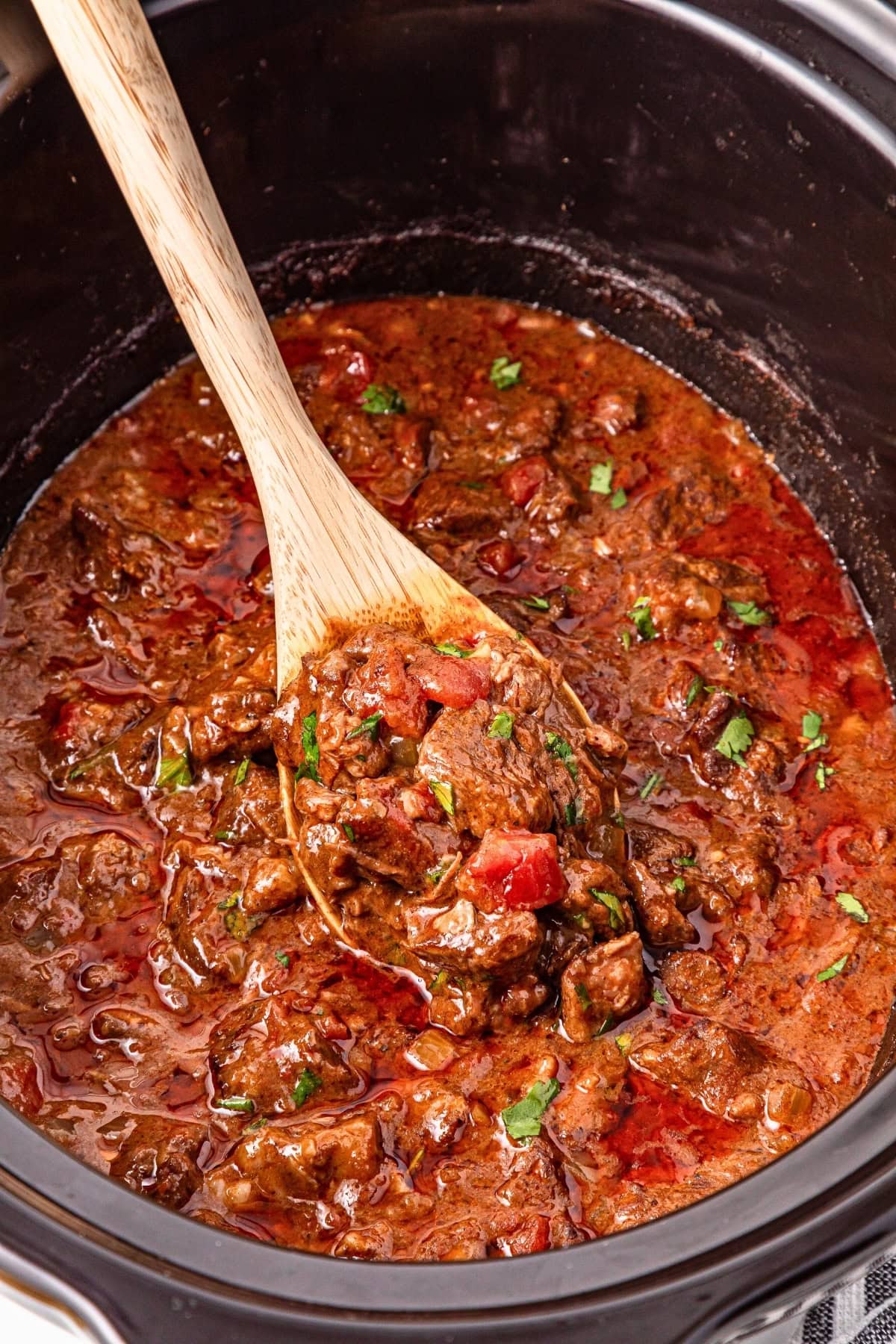 Beef stew with tomatoes in a slow cooker, stirred with a wooden spoon.