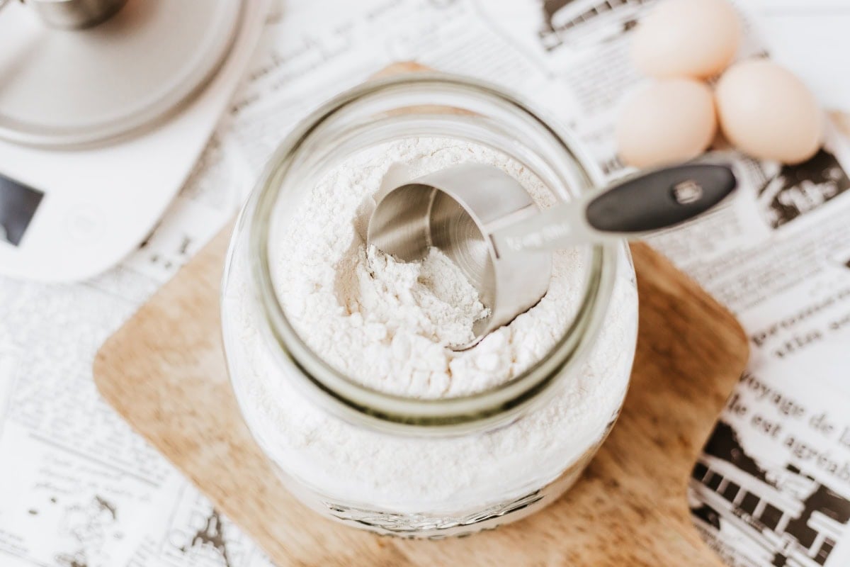 Jar of flour with a measuring cup on top, eggs in the background.
