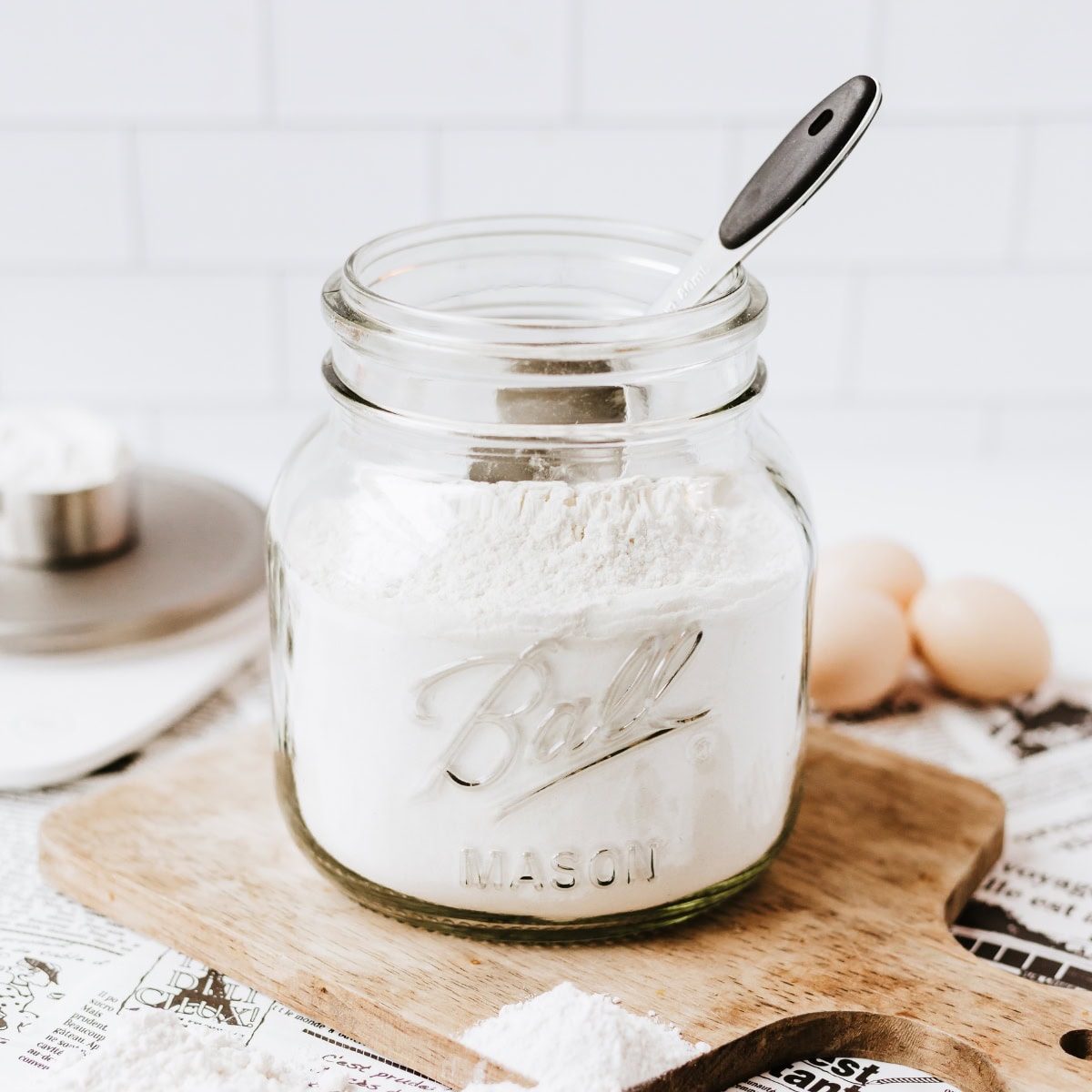 Glass jar of flour with a spoon on a wooden board, eggs in the background.