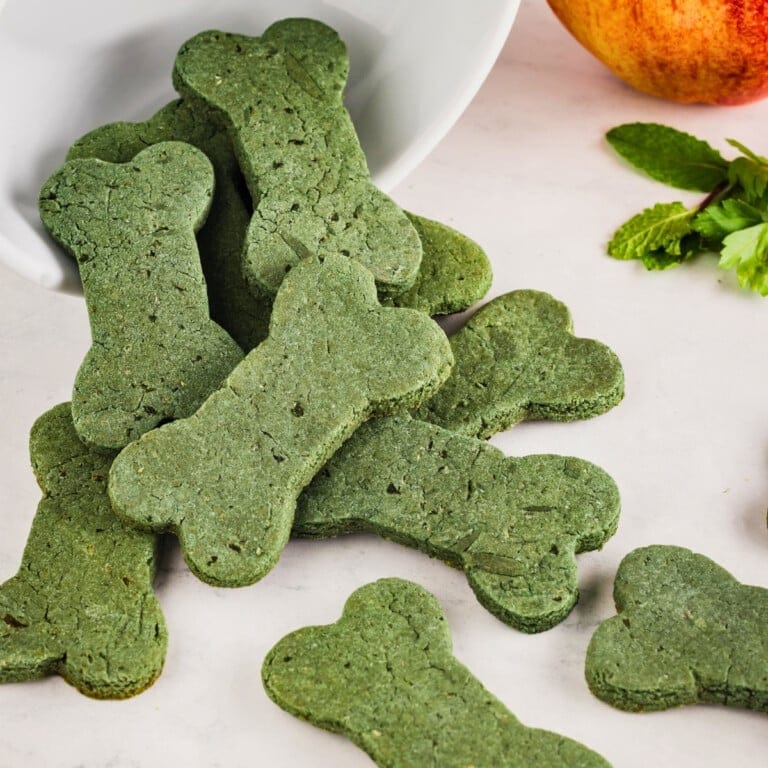 Green bone-shaped dog treats spilled from a white bowl onto a white surface.