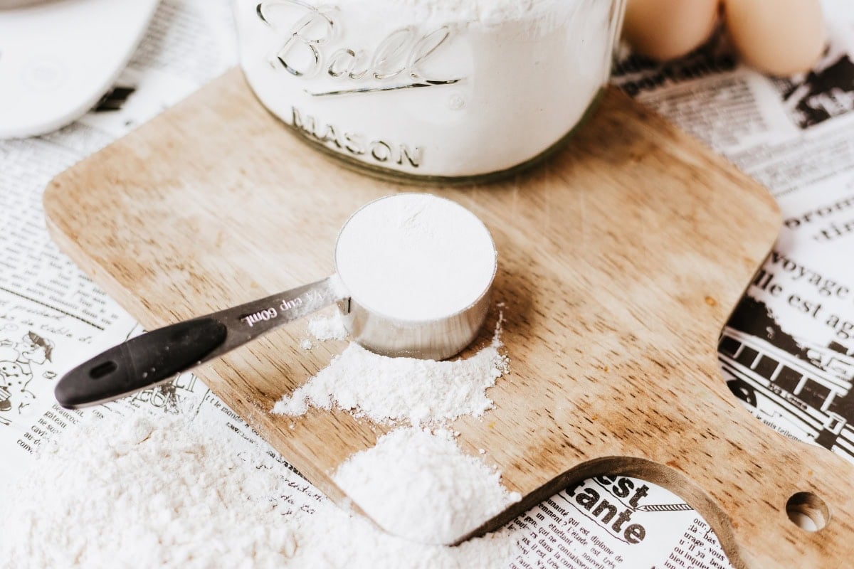 Measuring cup with flour on a wooden cutting board, newspaper underneath.
