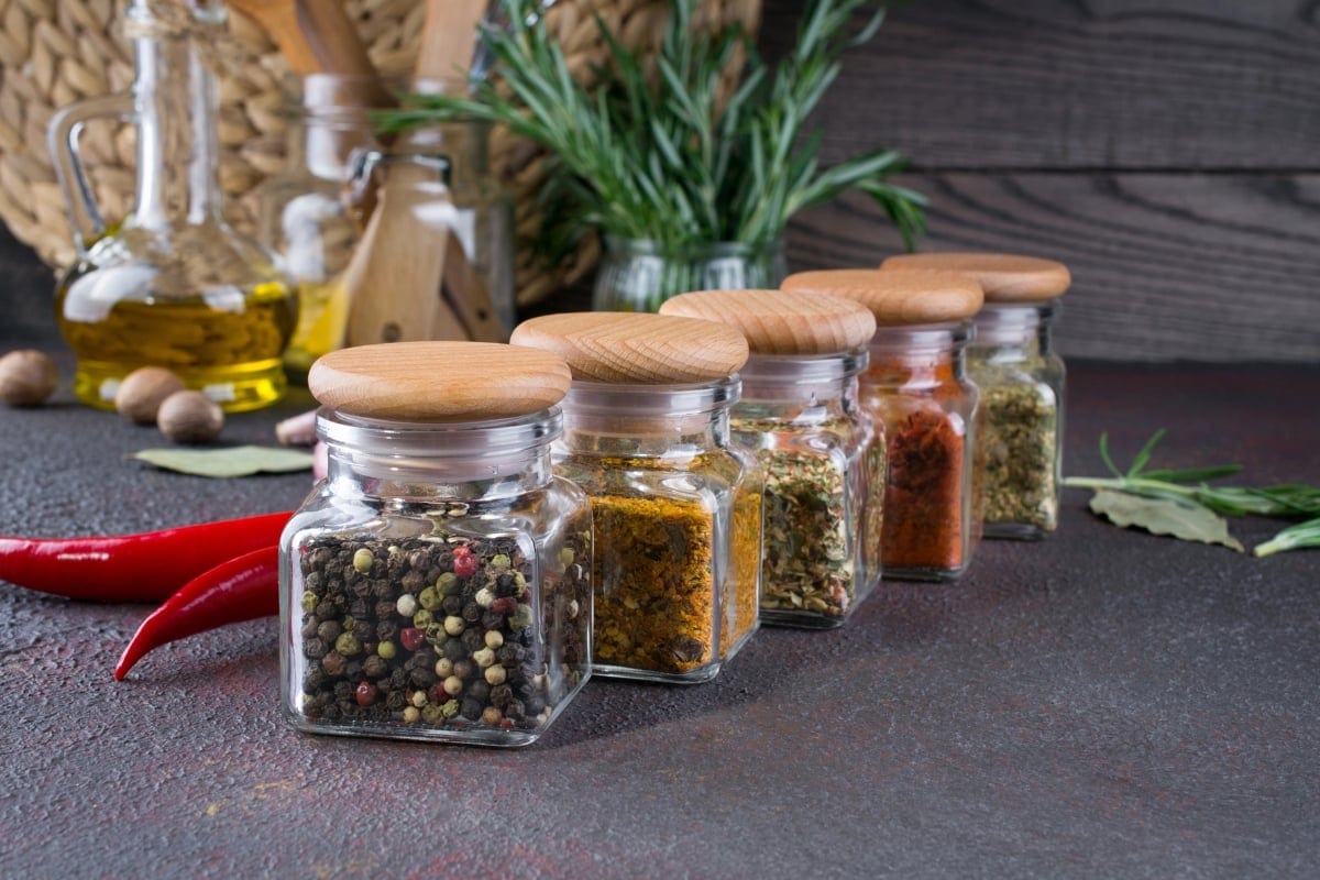 Spice jars with wooden lids on a counter, surrounded by herbs and oil.