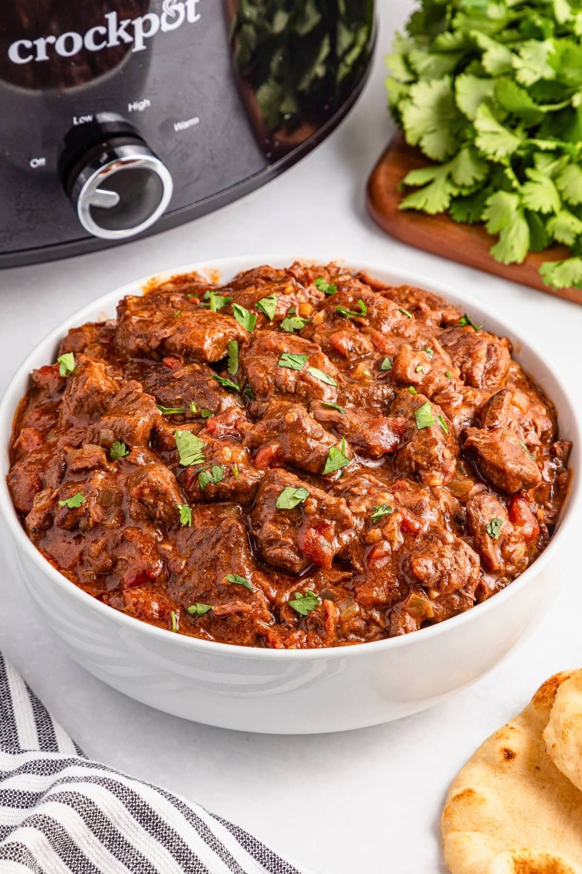 A bowl of beef stew garnished with herbs next to a Crockpot and fresh cilantro.