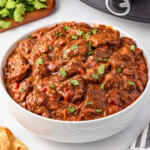 A bowl of beef stew garnished with chopped parsley on a white surface.