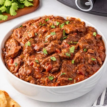A bowl of beef stew garnished with chopped parsley on a white surface.