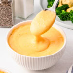 Spoon lifting creamy cheese sauce from a white bowl, with broccoli nearby.