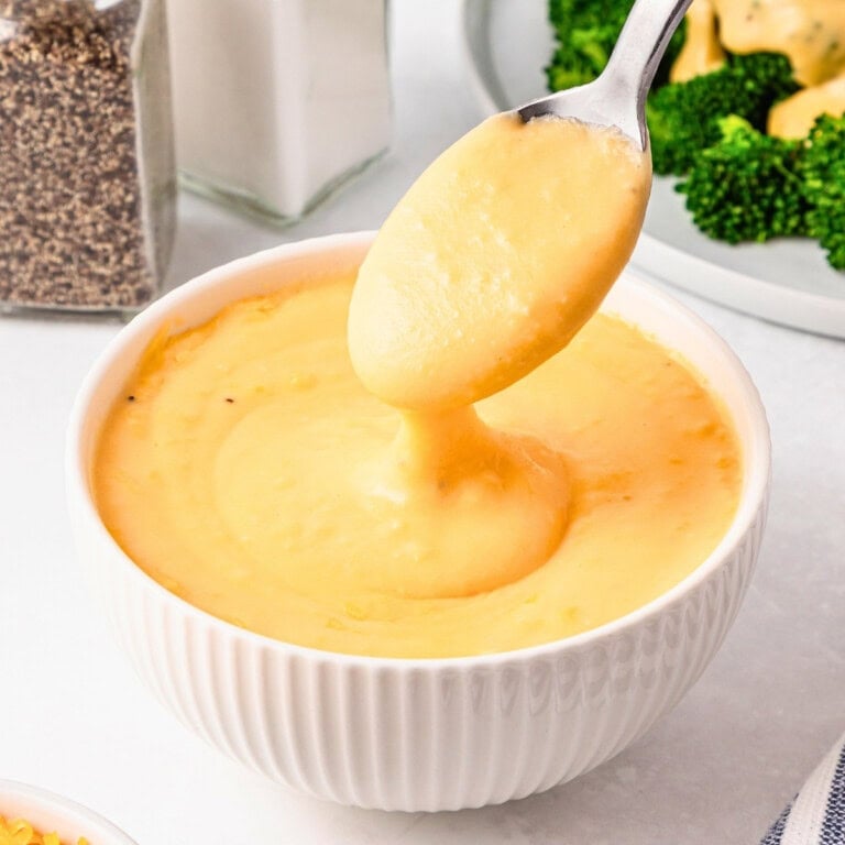 Spoon lifting creamy cheese sauce from a white bowl, with broccoli nearby.