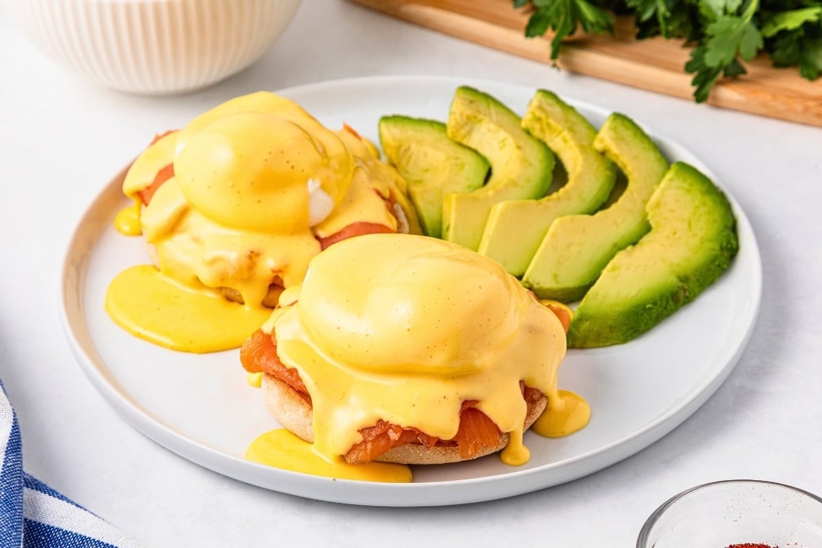 Eggs Benedict with hollandaise sauce and sliced avocado on a white plate.