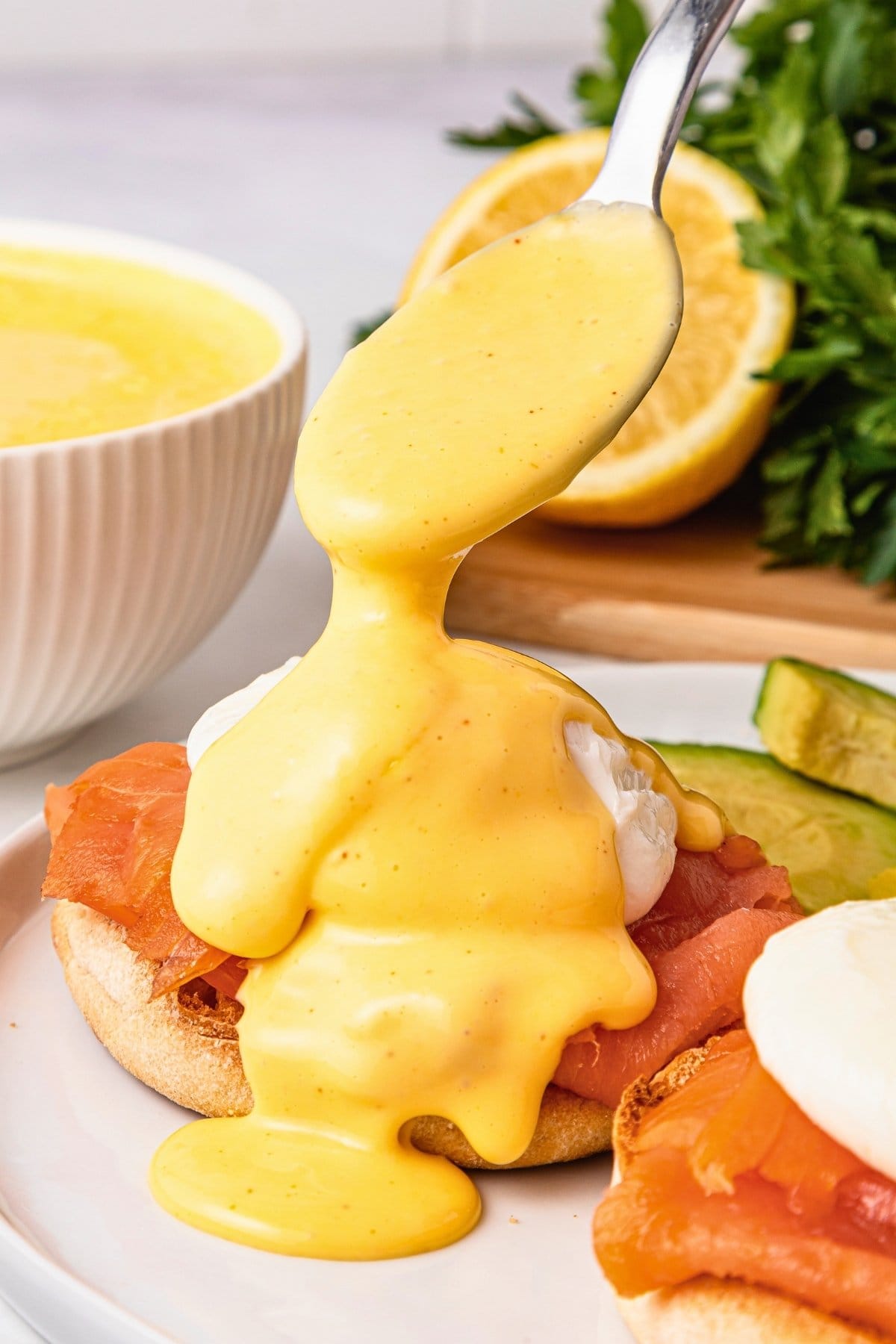 Spoon drizzling hollandaise sauce over eggs benedict with smoked salmon.