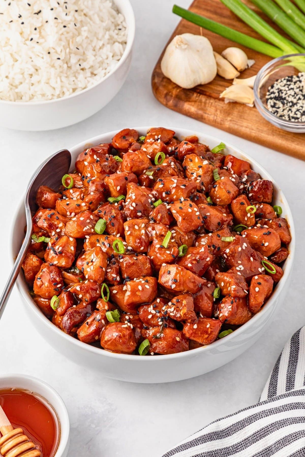 Bowl of sesame chicken garnished with green onions, with rice and ingredients nearby.