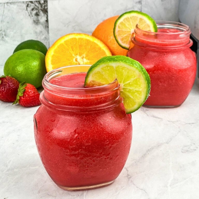 Two jars of strawberry slush topped with lime slices, with fruits in the background.