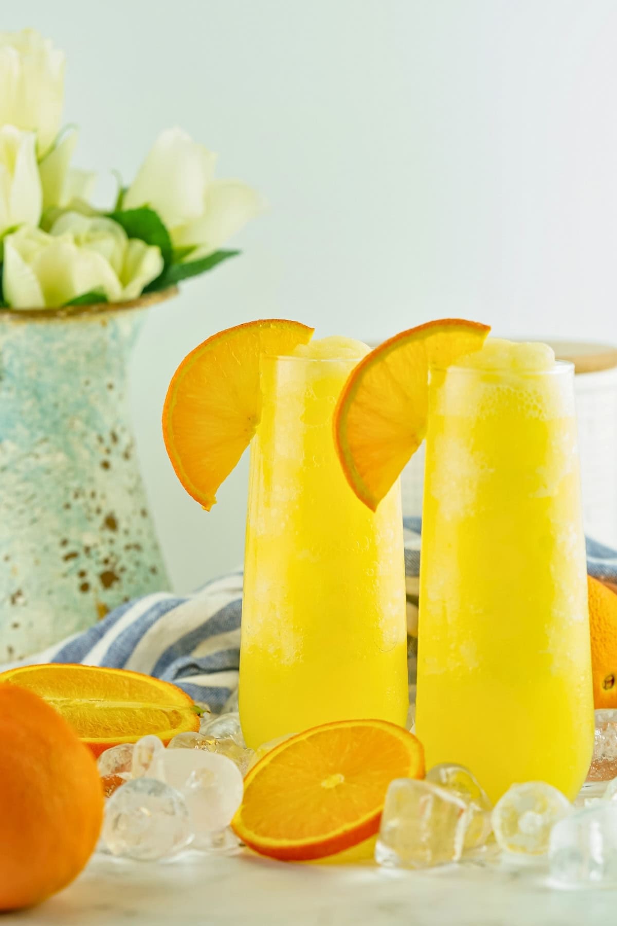 Two tall glasses of orange drink with orange slices, ice, and flowers in background.