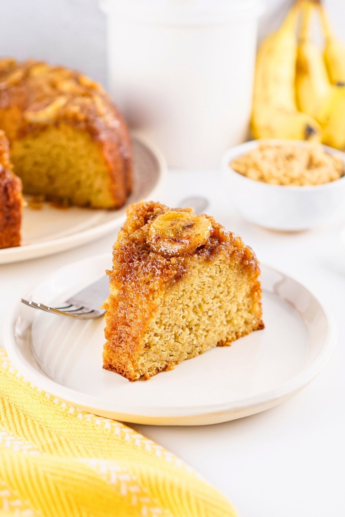 A slice of banana cake on a plate with a fork, bananas and cake in the background.