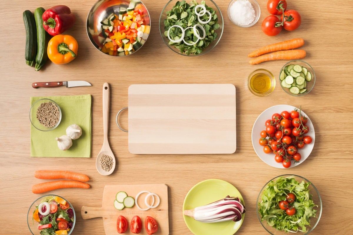 Chopping board surrounded by fresh vegetables, bowls, and utensils on a table.