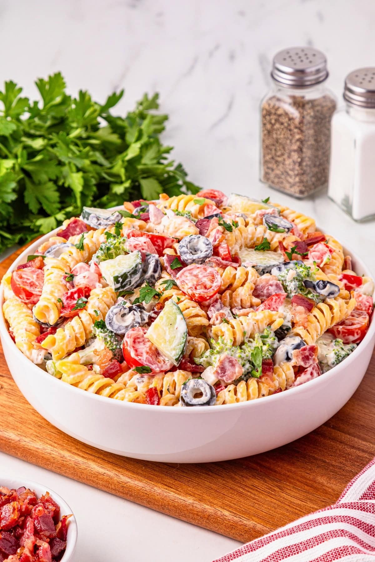 A bowl of creamy pasta salad with tomatoes, olives, and parsley on a wooden board.