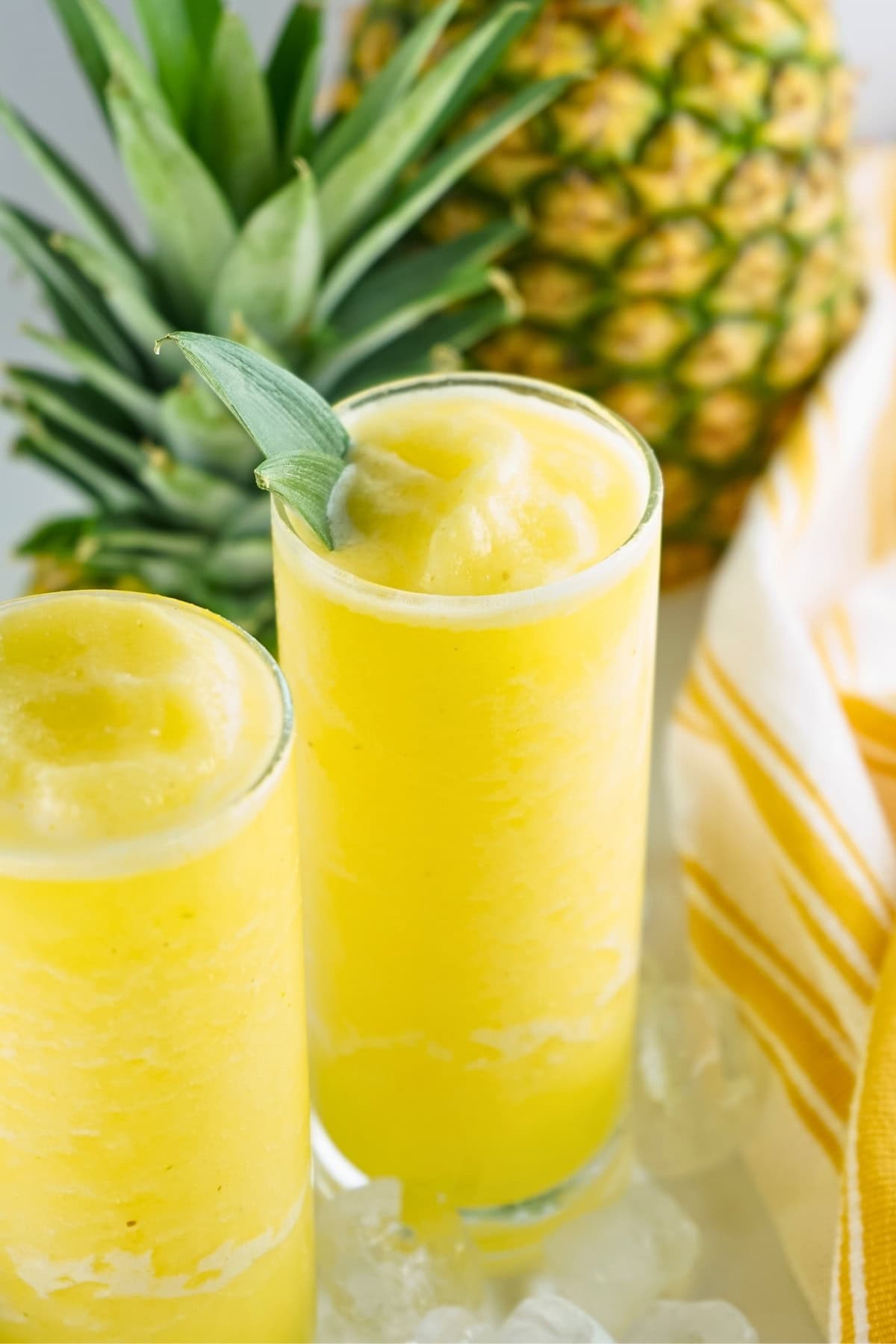 Two glasses of yellow frozen pineapple drink with a pineapple and striped towel nearby.