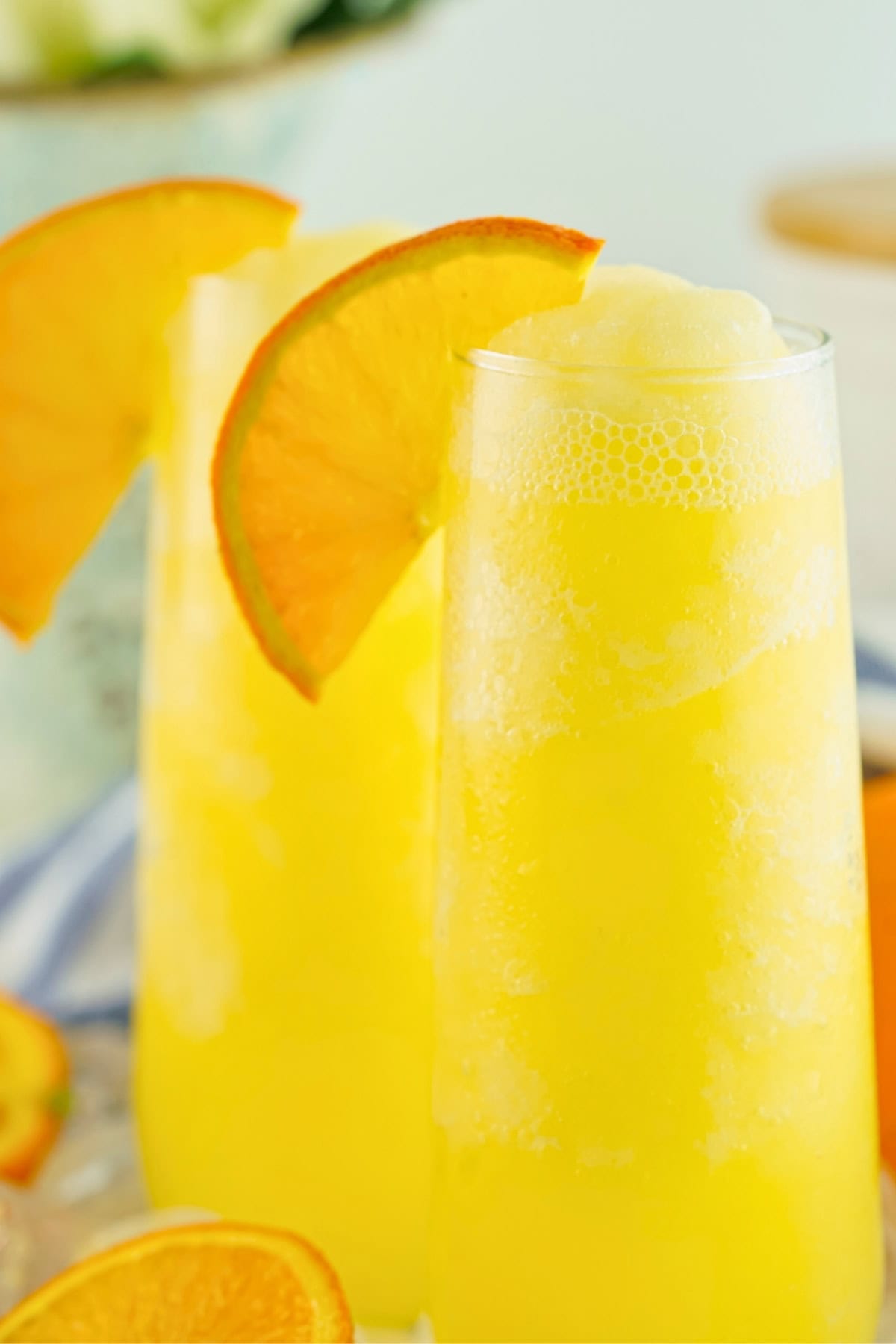 Two yellow frozen mimosas in tall glasses, garnished with orange slices.