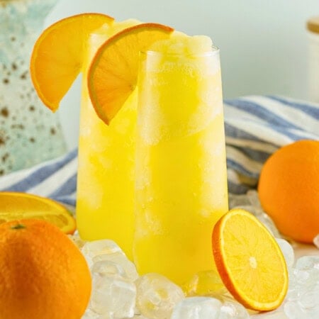 Two tall glasses of frozen mimosas with orange slices, surrounded by ice and oranges.