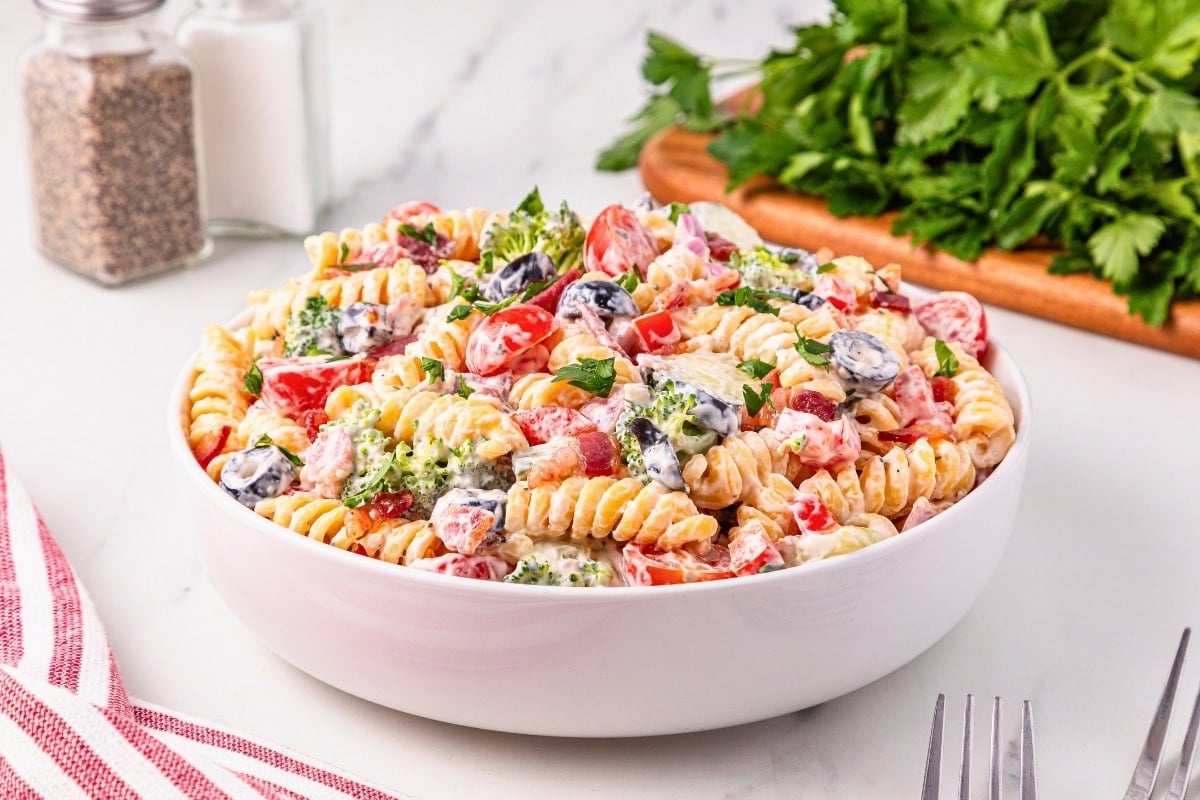 A bowl of creamy pasta salad with vegetables and herbs on a white table.