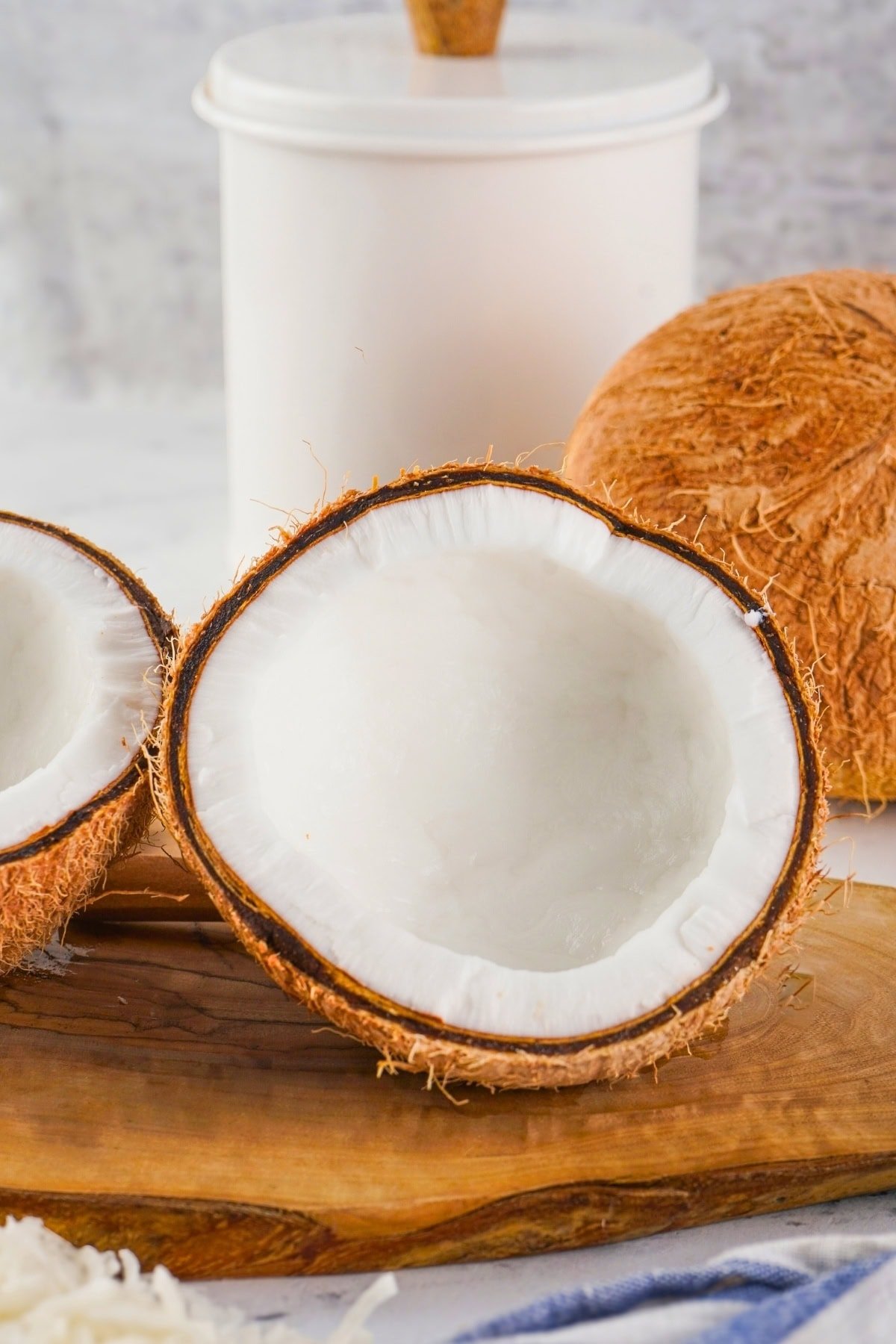 Halved coconut on a wooden board with whole coconut and white container behind.