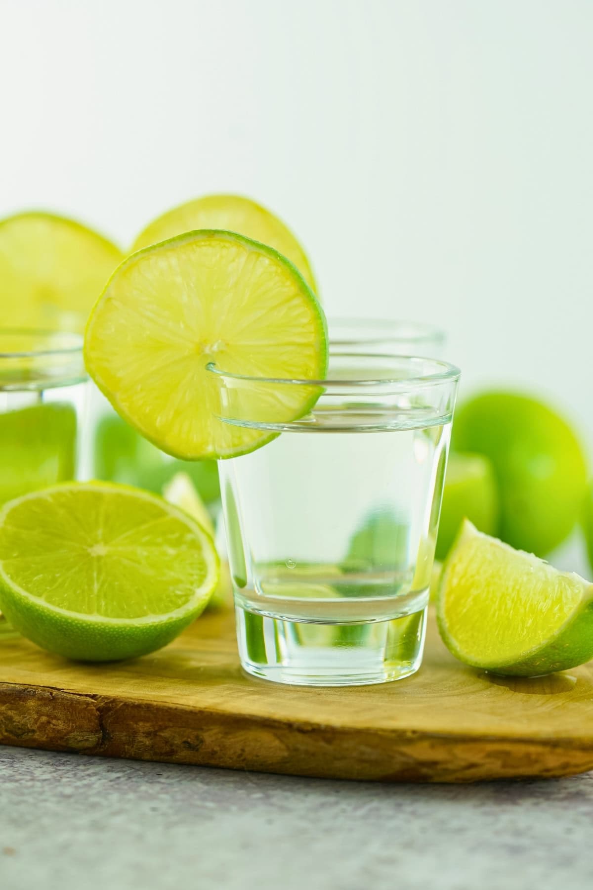 A shot glass with a lime slice, surrounded by cut limes on a wooden board.