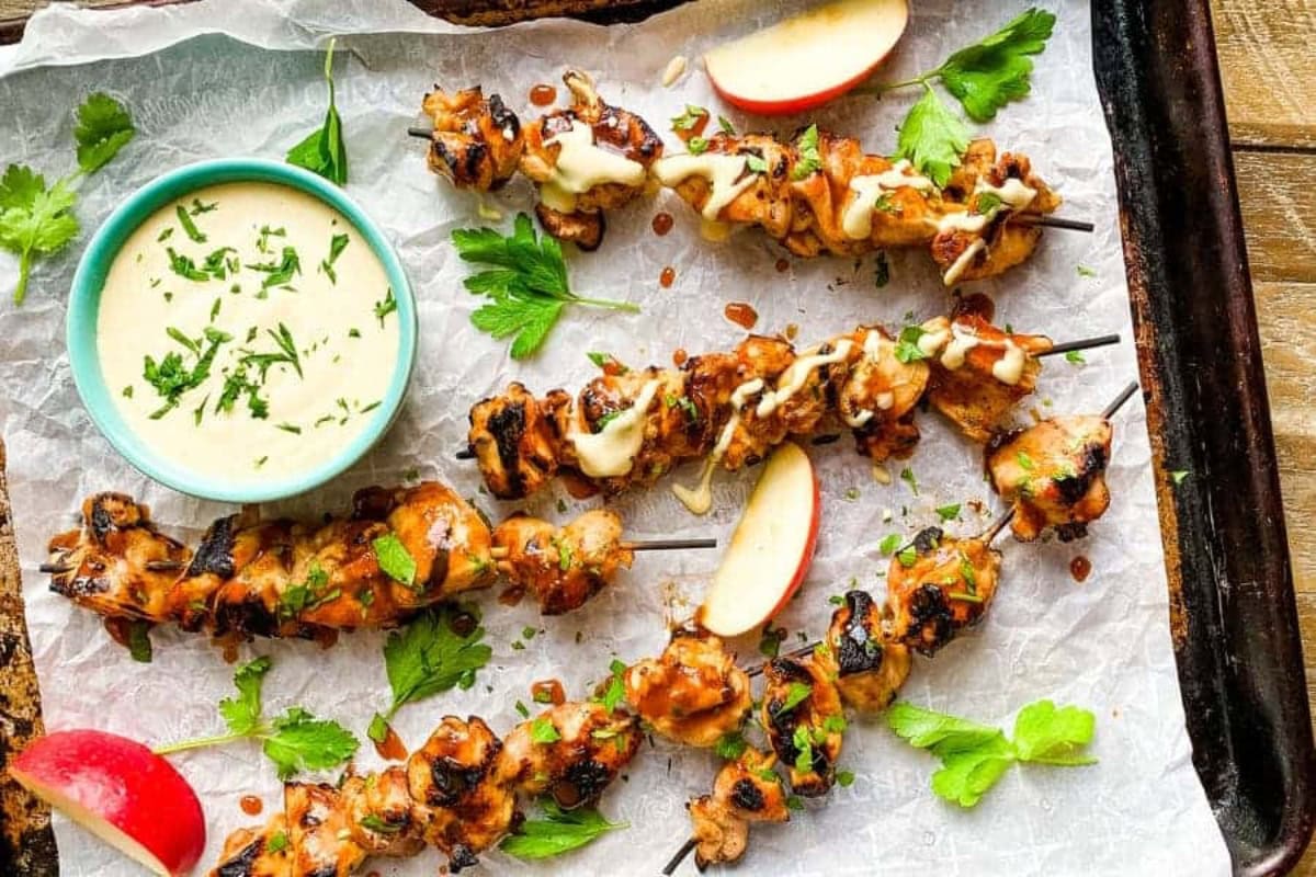 Grilled chicken skewers with sauce, parsley, and apple slices on parchment paper.