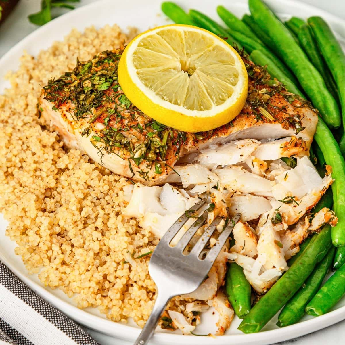 Seasoned fish fillet with lemon, quinoa, and green beans on a white plate.
