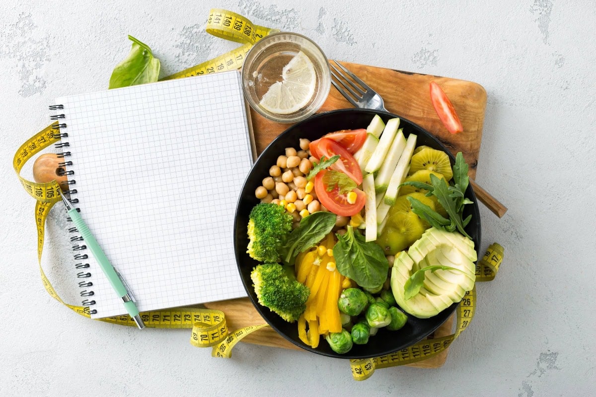A bowl of fresh veggies, a notepad, pen, tape measure, and lemon water.
