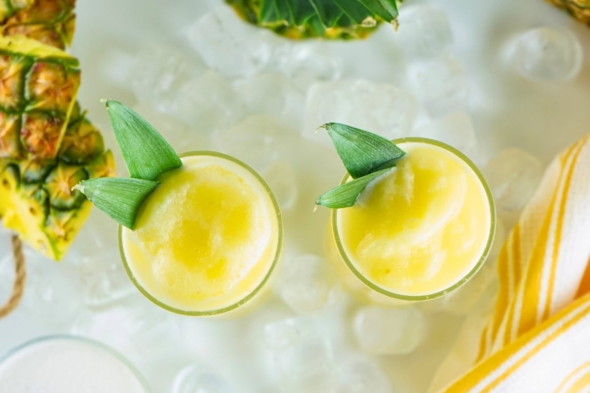 Two glasses of yellow frozen drinks with pineapple leaves, surrounded by ice.