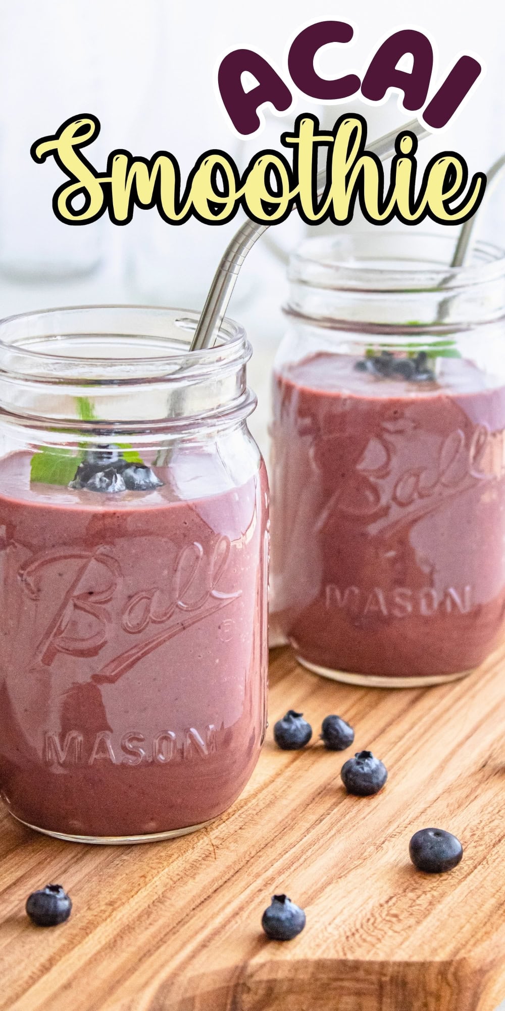 Two mason jars of acai smoothie with straws and fresh blueberries on a wooden board.