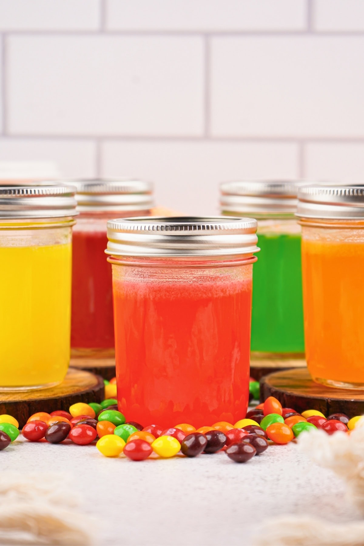 Four mason jars of colorful drinks with skittles in front of them.