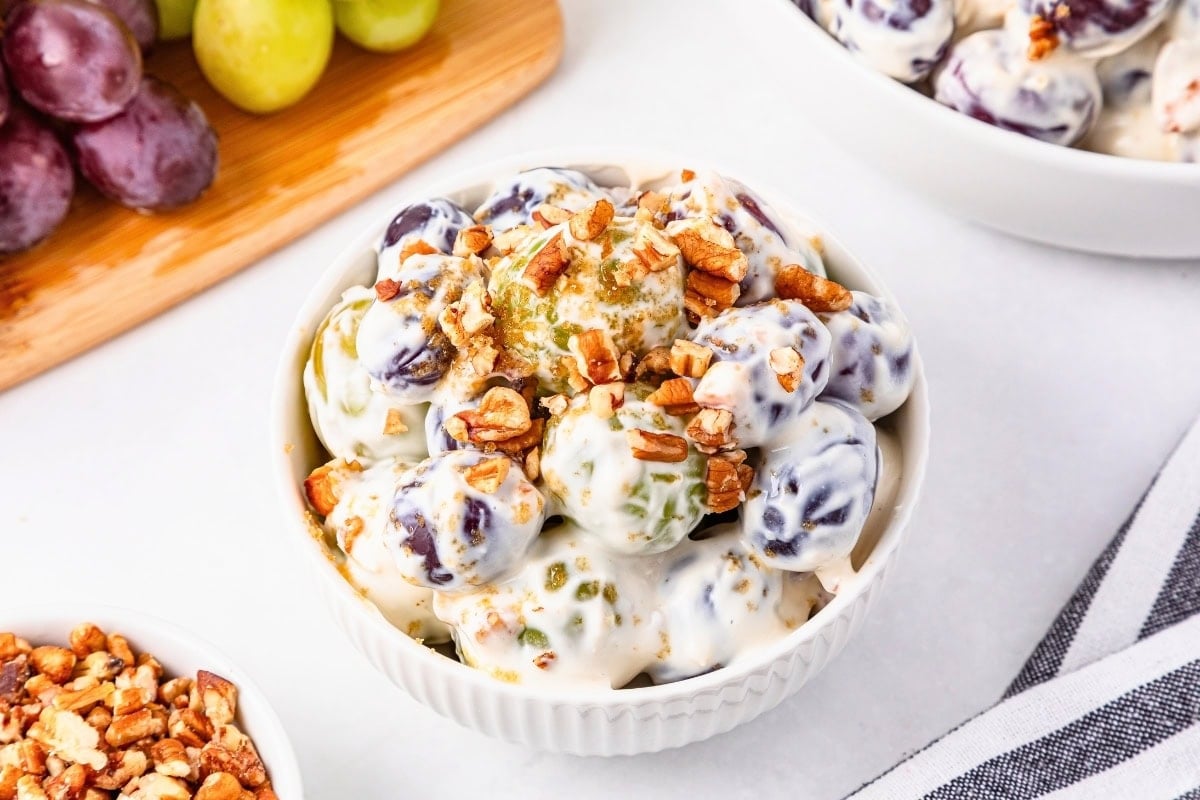 Bowl of grapes in creamy dressing topped with chopped pecans, with grapes nearby.