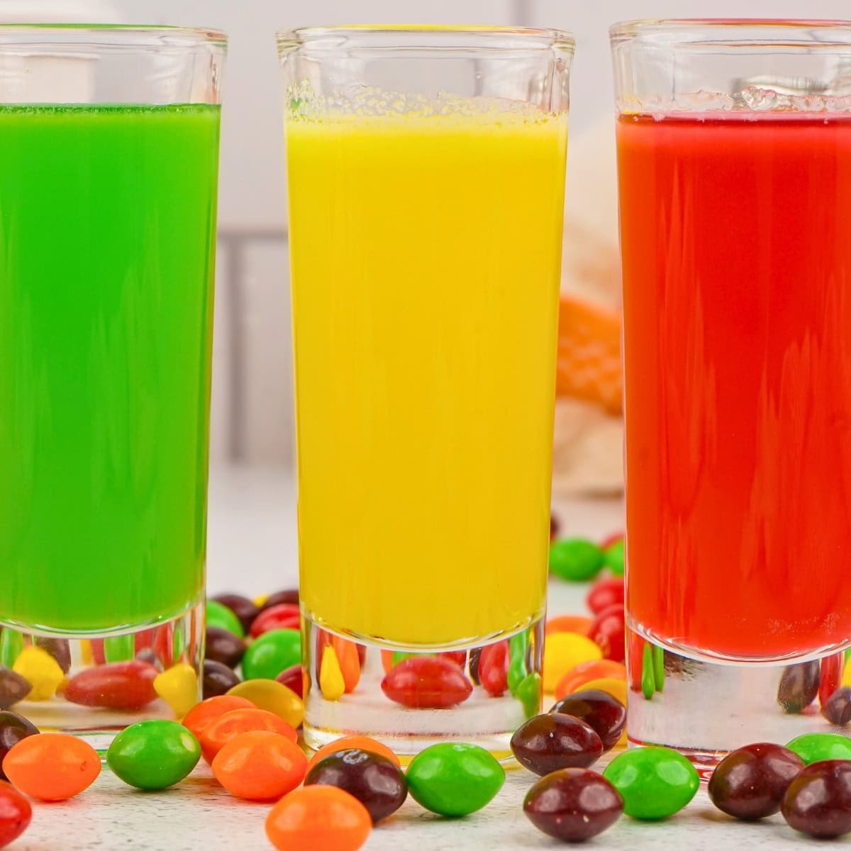 Three tall glasses of green, yellow, and red juice with colorful candies around them.