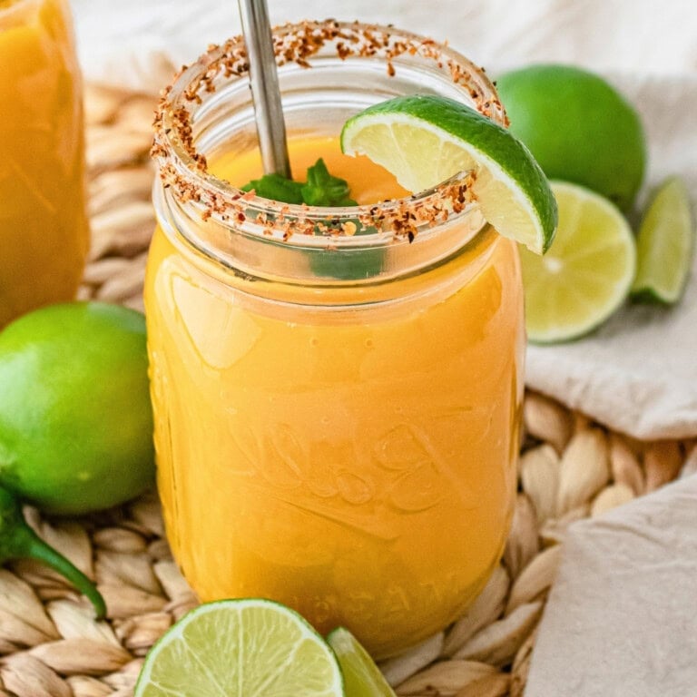 Mango drink in a mason jar with lime, chili rim, and metal straw on woven mat.