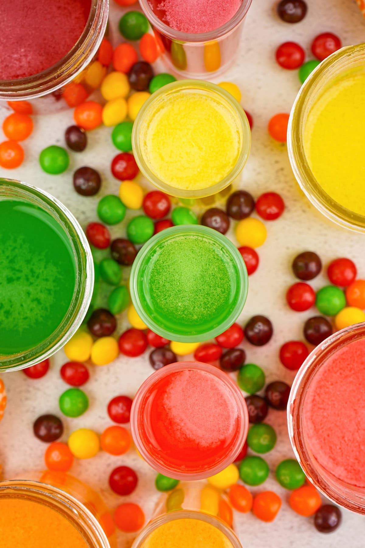 Colorful drinks in glasses surrounded by assorted skittle candies on a white surface.