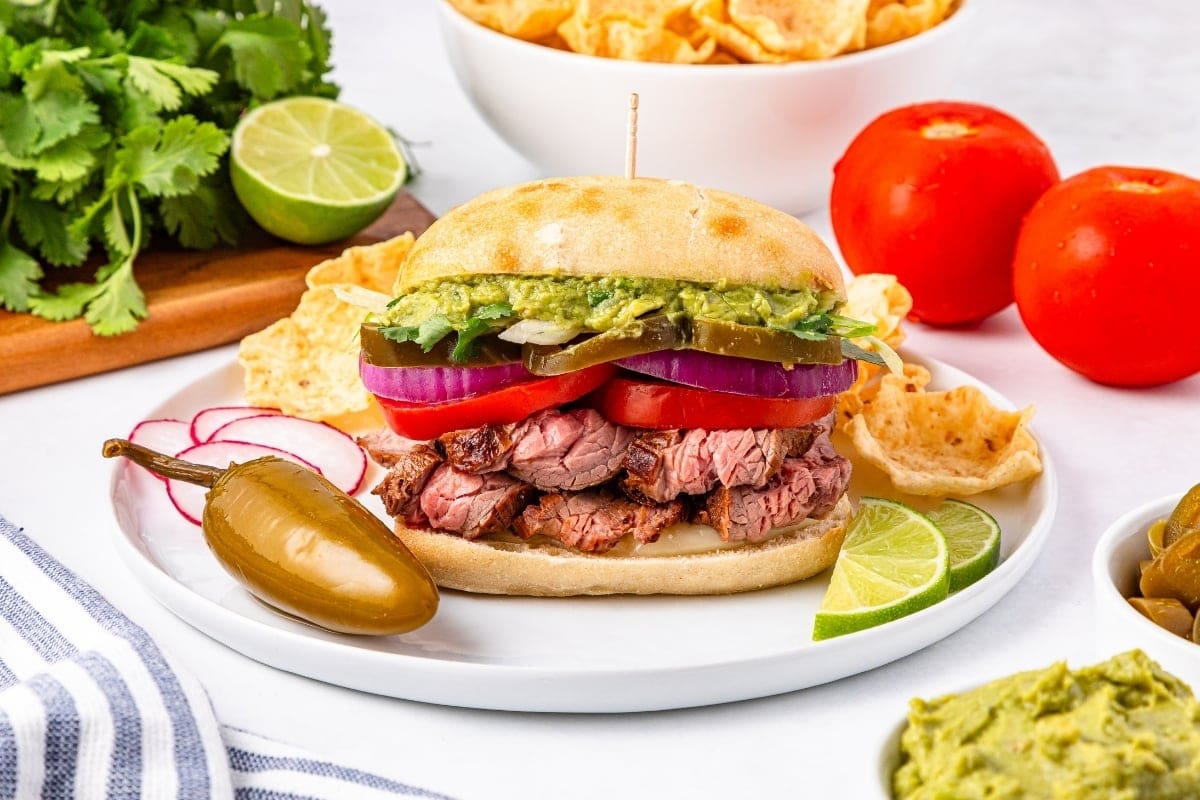 Sandwich with steak, guacamole, tomato, onions, and jalapeño on a plate.