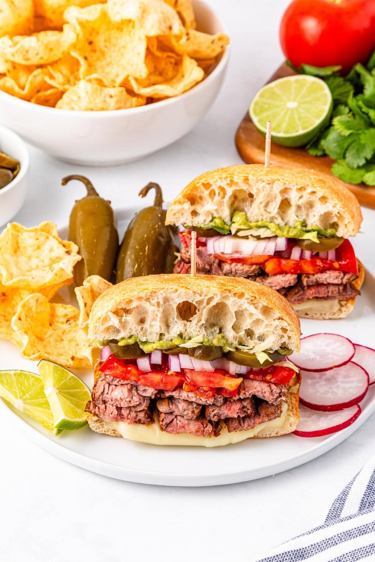 Steak sandwiches with veggies, chips, jalapeños, and lime on a white plate.