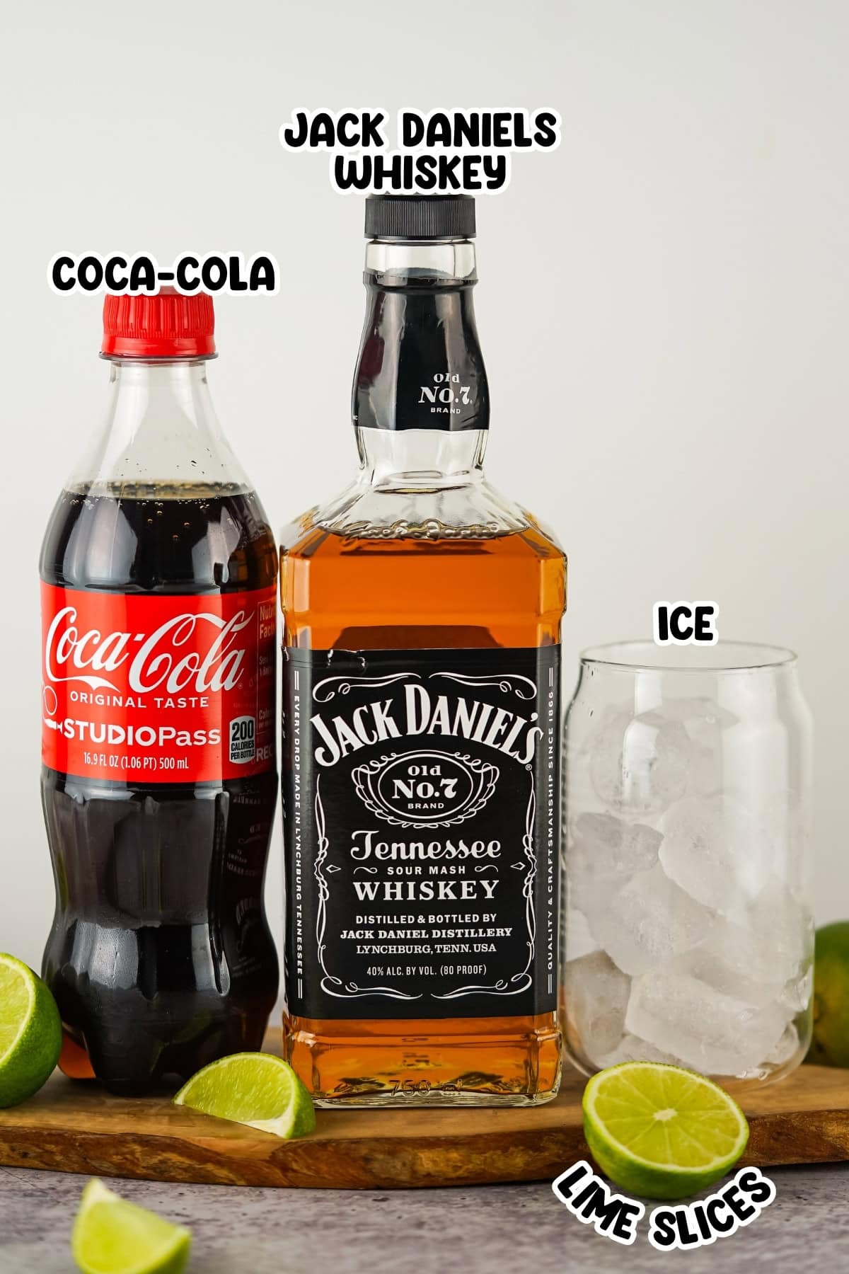 A bottle of Coca-Cola, Jack Daniels whiskey, a glass of ice, and lime slices.