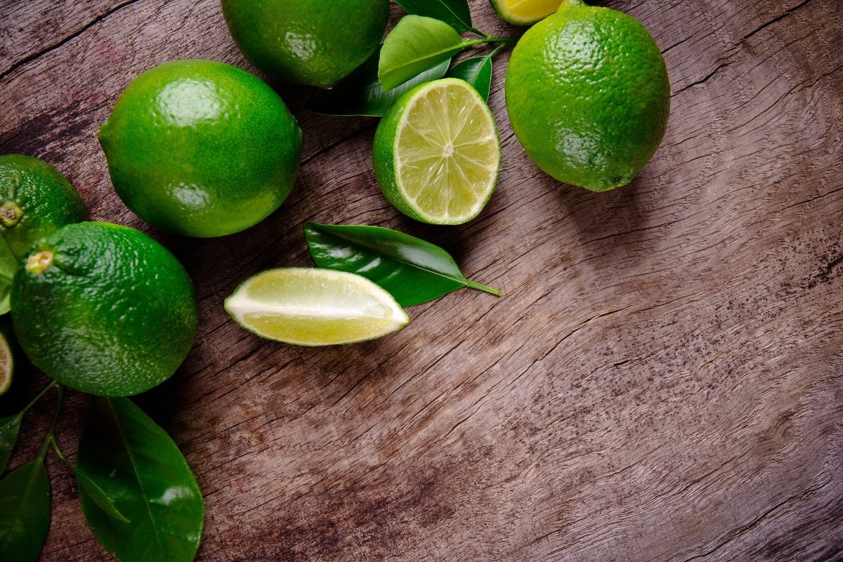 Whole and sliced limes with green leaves on a rustic wooden surface.