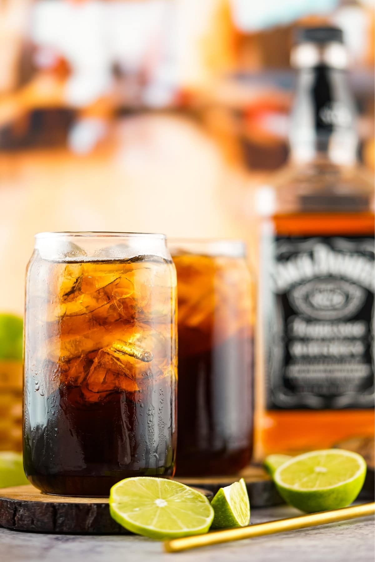 Two glasses of cola with ice and lime, with a whiskey bottle in the background.