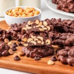 Chocolate clusters with peanuts stacked on a wooden board, bowl of peanuts behind.