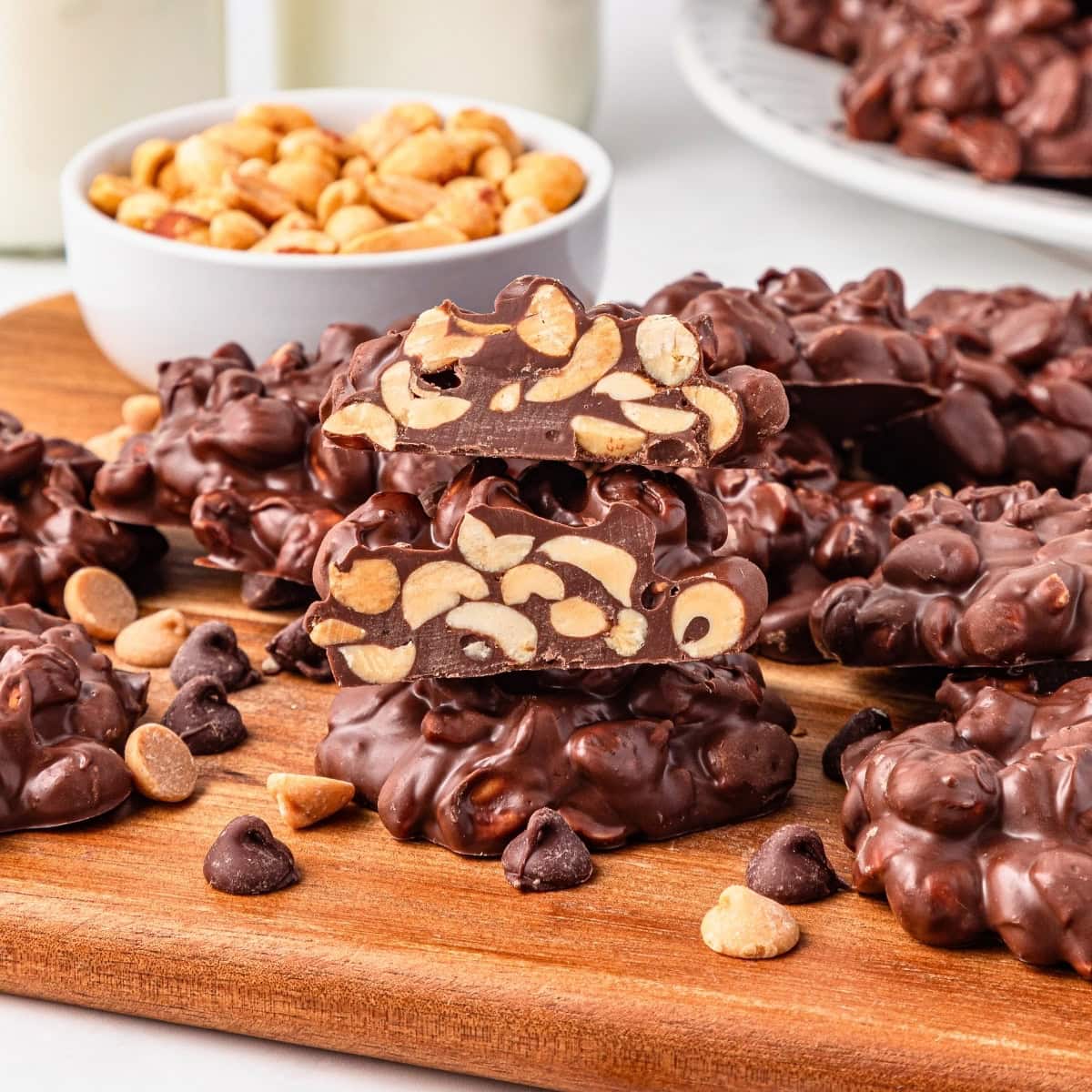 Chocolate clusters with peanuts stacked on a wooden board, bowl of peanuts behind.