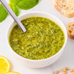 Bowl of green pesto with a spoon, surrounded by bread, basil, and lemon slices.