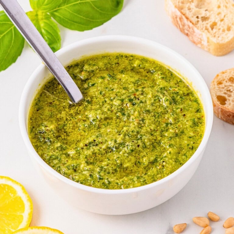 Bowl of green pesto with a spoon, surrounded by bread, basil, and lemon slices.